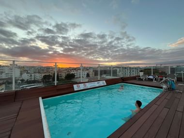 Dachpool im Hotel Marina Rio in Lagos mit zwei Schwimmern bei Sonnenuntergang. Dramatischer orange-blauer Himmel über der Stadtsilhouette.