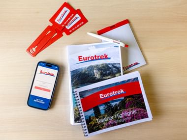 Holztisch mit Eurotrek-Reiseunterlagen, Broschüren mit Berglandschaften, roten Gepäckanhängern und Smartphone mit Eurotrek-Website.