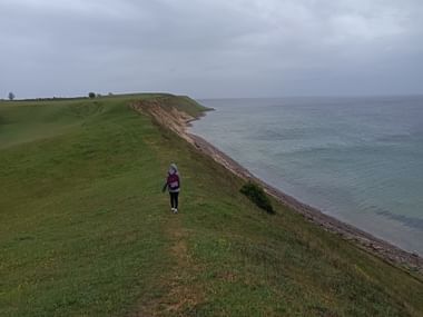 Wanderin auf grüner Küstenklippe am Österlenleden zwischen Ystad und Kåseberga, mit ruhigem Meer und bewölktem Himmel.