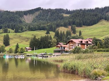 Hostellerie Schwarzsee, ein Holzhotel mit rotem Dach, neben einem See mit Schilf. Grüne Wiesen und bewaldete Hügel im Hintergrund.