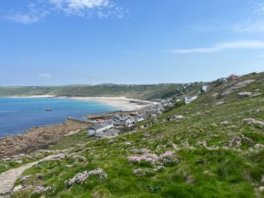 Küstendorf mit weißen Häusern am Hang mit Blick auf blaue Bucht und Sandstrand in Cornwall. Rosa Wildblumen blühen auf grünen Hängen.