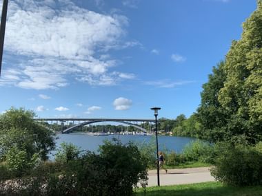 Blick auf eine hohe Bogenbrücke über blaues Wasser mit grünen Bäumen am Ufer und einem Radfahrer auf einem Uferweg unter blauem Himmel.