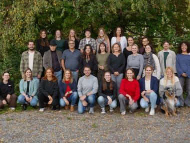 Gruppe von etwa 25 Personen in drei Reihen für ein Teamfoto im Freien arrangiert, mit grünem Laubhintergrund und einem Hund in der ersten Reihe.