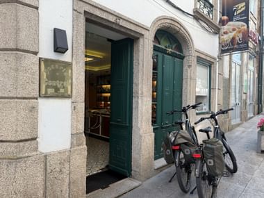 Zwei E-Bikes mit Taschen vor dem Geschäft Charutos dos Arcos in Arcos de Valdevez. Grüne Holztüren rahmen den Eingang.