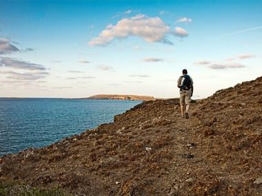 Minorque attire par ses sentiers côtiers pittoresques. Randonneur avec sac à dos marchant sur sentier côtier rocheux à Minorque avec mer bleue et île distante sous ciel nuageux.