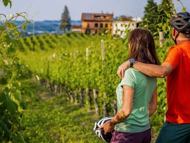 Deux cyclistes avec casques debout parmi les rangées de vignes vertes au Piémont, Italie, avec des bâtiments traditionnels visibles.