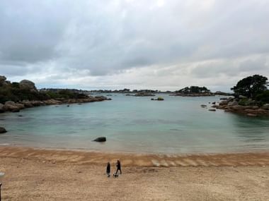 Zwei Personen am Sandstrand mit Blick auf türkisfarbene Bucht mit rosa Granitfelsen und grüner Vegetation unter bewölktem Himmel in Ploumanac'h.