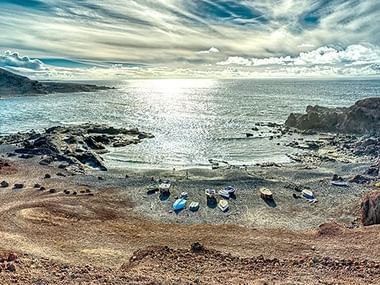 Küstenbucht auf Lanzarote mit kleinen Fischerbooten am Sandstrand, umgeben von vulkanischen Felsformationen unter dramatischem Himmel.