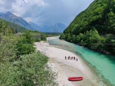 Fluss Soca in Slowenien. Mehrere Personen und ein rotes Raftingboot sind am Ufer. Umgeben von Bäumen und Bergen.