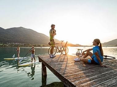 Deux cyclistes sur ponton en bois au Fuschlsee au coucher du soleil, avec des pratiquants de paddle sur le lac et montagnes en arrière-plan.