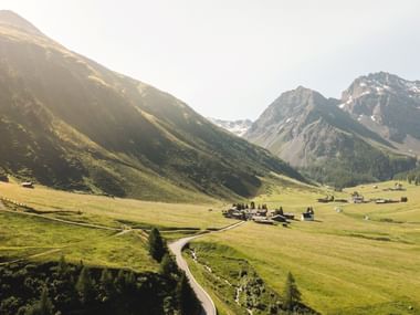Luftaufnahme des Dorfes Sertig im Sertigtal, umgeben von grünen Almwiesen und schneebedeckten Bergen unter goldenem Sonnenlicht.