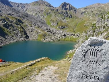 Marqueur en pierre indiquant 'Quelle Rhein' près d'un lac alpin turquoise entouré de montagnes rocheuses. Une tente rouge est visible sur la rive.