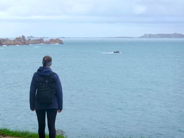 Frau mit Rucksack betrachtet die bretonische Küste bei Ploumanac'h. Rosa Granitfelsen links, Leuchtturm auf ferner Insel rechts.