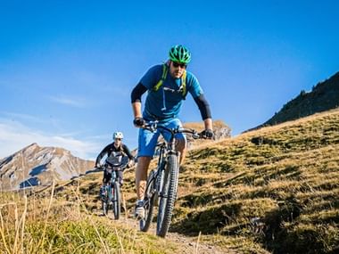 Les vététistes en descente vers la vallée. Deux vététistes descendent une pente alpine herbeuse en Suisse centrale, avec des montagnes enneigées en arrière-plan.