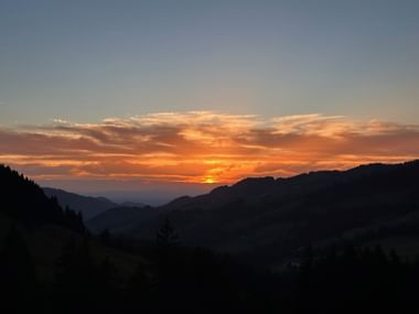 Sonnenuntergang über bewaldeten Bergkämmen bei der Gantrischhütte. Orange und goldene Wolken erhellen den Himmel über dunklen Hügeln.