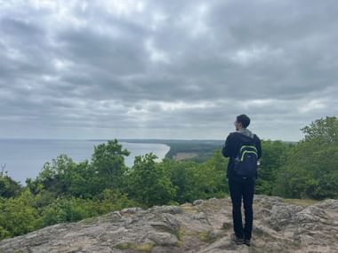 Wanderer mit Rucksack auf felsigem Aussichtspunkt mit Blick auf grüne Küste und Meer unter bewölktem Himmel am Österlenleden bei Kivik.