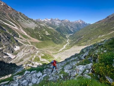 Randonneur avec sac à dos rouge descendant un terrain rocheux surplombant la vaste vallée du Pitztal avec des sommets alpins enneigés.