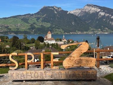 Holzschild Spiez mit Seeblick, historischem Schloss mit Turm, Bergen und Thunersee. Text: 'Kraftort am Wasser'.