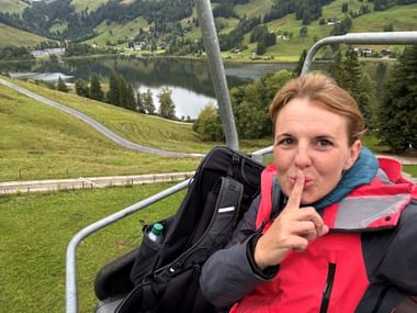 Frau in roter Jacke im Sessellift zur Riggisalp, mit dem Schwarzsee und grünen Almwiesen im Hintergrund.