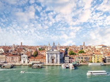 Panoramic view of Venice waterfront showing historic buildings with red tile roofs, a prominent white church, and boats on turquoise water.