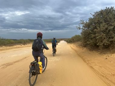 Zwei Radfahrer auf einem Sandweg bei Lagoa, Algarve. Der Weg ist von Vegetation gesäumt unter bewölktem Himmel.