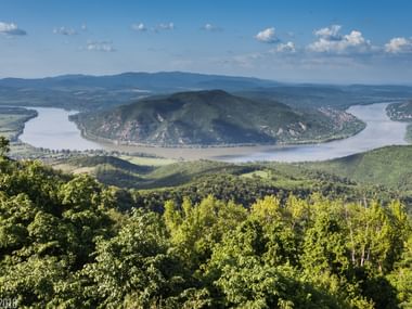 Luftaufnahme der Donau, die eine Hufeisenbiegung um einen bewaldeten Hügel bildet, mit grünen Tälern und fernen Bergen unter blauem Himmel.