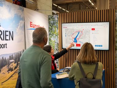 Drei Besucher am Eurotrek-Stand betrachten digitale Karte auf Bildschirm. Mitarbeiterin zeigt auf Route mit Berglandschaft im Hintergrund.
