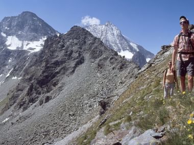 Two hikers on the Alpine Pass Trail in Valais with a view of the rocky mountain landscape in the background.