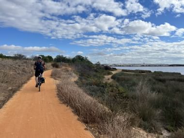 Radfahrer auf orangefarbenem Schotterweg zwischen Lagos und Portimão entlang der Algarve-Küste mit Lagune, trockener Vegetation und blauem Himmel.