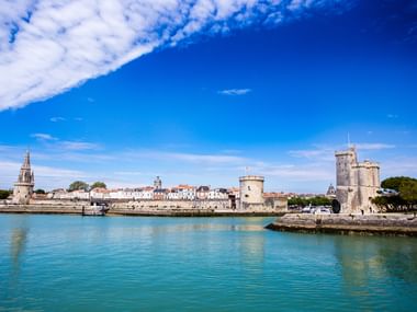 Port historique sur la côte atlantique avec tours de pierre médiévales et fortifications reflétées dans l'eau turquoise sous ciel bleu.