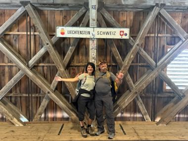Zwei Wanderer mit Rucksäcken posieren unter einem Liechtenstein-Schweiz-Grenzschild auf einer überdachten Holzbrücke mit diagonalen Balken.