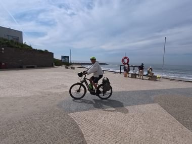 Radfahrer mit gelbem Helm und Packtaschen auf gepflasterter Küstenpromenade. Ozean und Strand rechts sichtbar, Menschen im Hintergrund am Wasser.