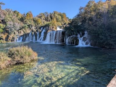 Breite Wasserfälle stürzen in klaren türkisfarbenen Pool in Norddalmatien, umgeben von Herbstbäumen unter blauem Himmel.