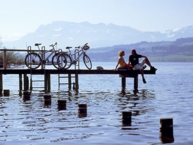 VELO Zwei Radfahrer sitzen auf Holzsteg mit Fahrrädern und blicken auf Schweizer See mit Bergen im Hintergrund. Holzpfosten im Wasser sichtbar.