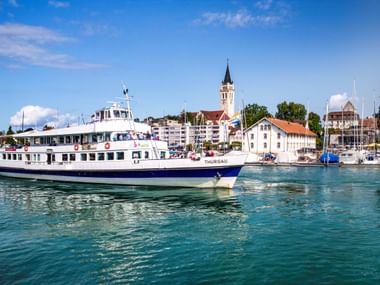 Weißes Passagierboot namens Thurgau im Romanshorner Hafen mit Kirchturm und historischen Gebäuden im Hintergrund unter blauem Himmel.