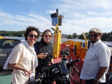 Drei Radfahrer mit Fahrrädern auf gelbem Fährdeck in Stockholm. Wasser und grünes Ufer im Hintergrund unter blauem Himmel sichtbar.