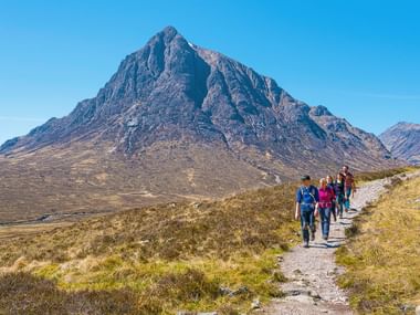Gruppe von Wanderern auf Steinpfad durch schottische Highlands mit dramatischem Buachaille Etive Mor Gipfel im Hintergrund unter blauem Himmel.