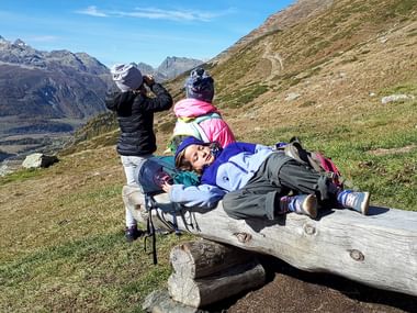 Drei Kinder machen Pause. Ein Mädchen liegt auf einem Holzbank, die anderen zwei schauen sich die Berge an.