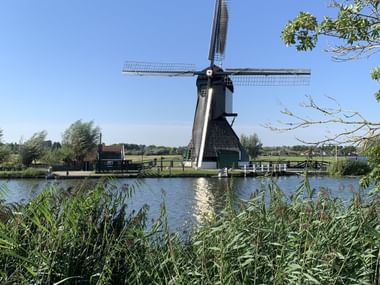 Man sieht eine dunkle Mühle hinter einem Kanal und viel Grün.