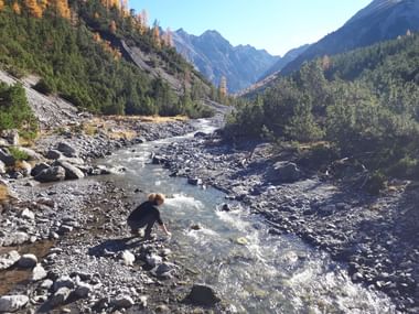 Frau kniet an einem steinigen Bergbach im Schweizer Nationalpark-Tal, umgeben von Herbstwald und schneebedeckten Gipfeln.