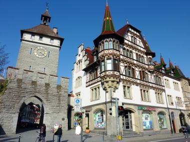 Mittelalterlicher Steinturm mit Uhr und verziertes Fachwerkgebäude in der Konstanzer Altstadt. Klarer blauer Himmel und Fußgänger auf der Straße.