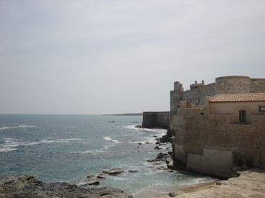 Küstenfestung im Südosten Siziliens Steinfestung und Gebäude direkt an der Mittelmeerküste im Südosten Siziliens mit türkisfarbenem Wasser und felsiger Küstenlinie.