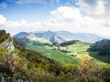 Jura Gebirge Panoramablick ins Tal Panoramablick auf die Schweizer Jura-Berge mit grünen Hügeln, verstreuten Bauernhöfen und bewaldeten Hängen unter bewölktem Himmel.