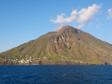 Blick vom Meer auf eine Vulkaninsel mit grünen Hängen und weißen Häusern entlang der Küste unter blauem Himmel mit weißen Wolken.
