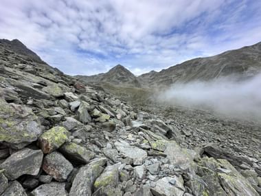 Steiniger Wanderweg am Alpenpässeweg mit grauen, moosbedeckten Felsen, umgeben von Berggipfeln und tiefliegendem Nebel.