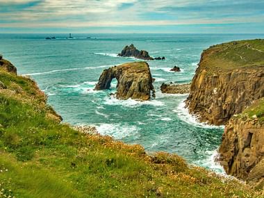 Côte dramatique de Cornouailles avec falaises vertes, mer turquoise et formation d'arche rocheuse. Vagues blanches contre falaises dorées.