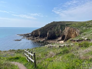 Küstenwanderweg mit weißem Holzzaun in Cornwall, mit Blick auf felsige Klippen und blaues Meer unter klarem Himmel.