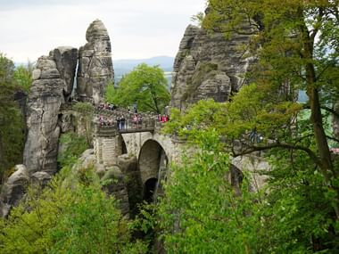 Die historische Basteibrücke erstreckt sich zwischen hohen Sandsteinfelsen, umgeben von grünem Wald, mit Besuchern auf der Steinbogenbrücke.