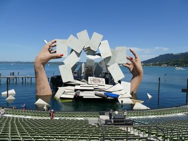 Seebühne Bregenz schwimmende Bühne mit riesigen Händen die weiße Karten halten, Amphitheater und Bodensee mit Bergen im Hintergrund.