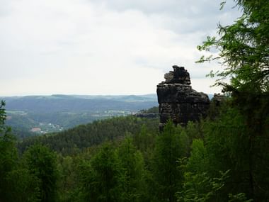 Markante Felsformation über dichtem Wald an der Riedelaussicht, mit sanften Hügeln und Dörfern in der Ferne.